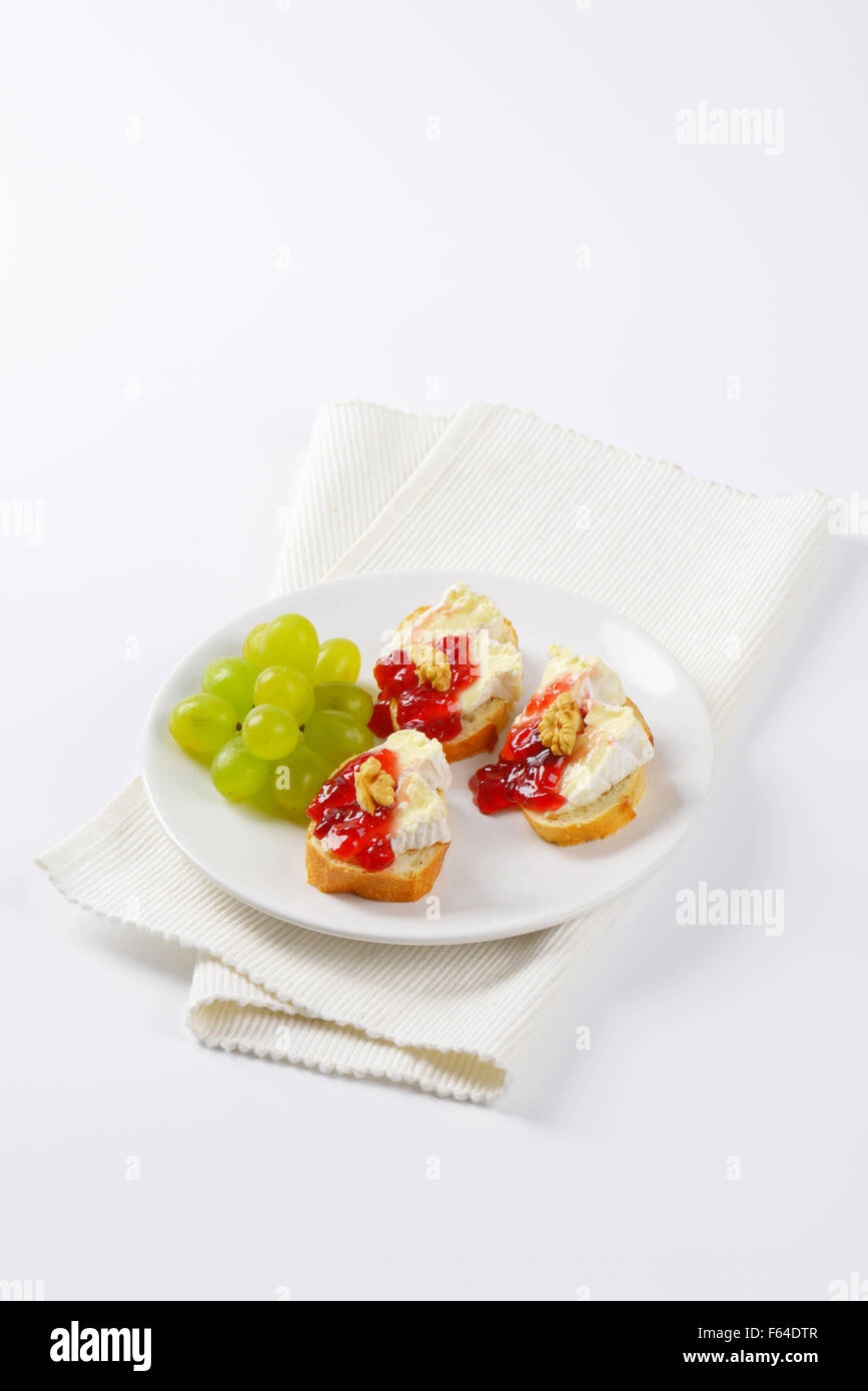slices of fresh bread with brie cheese, walnuts, jam and grapes on white plate Stock Photo