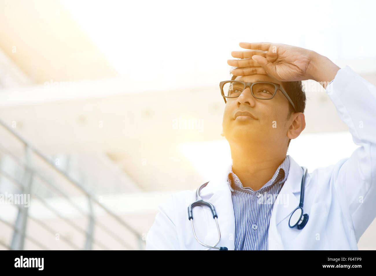 Portrait of Asian Indian medical doctor hand shielded looking away, standing outside hospital building, beautiful golden sunligh Stock Photo