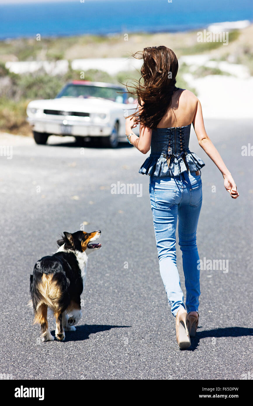 Rear view of woman with windswept hair wearing jeans and top walking with dog on street Stock Photo