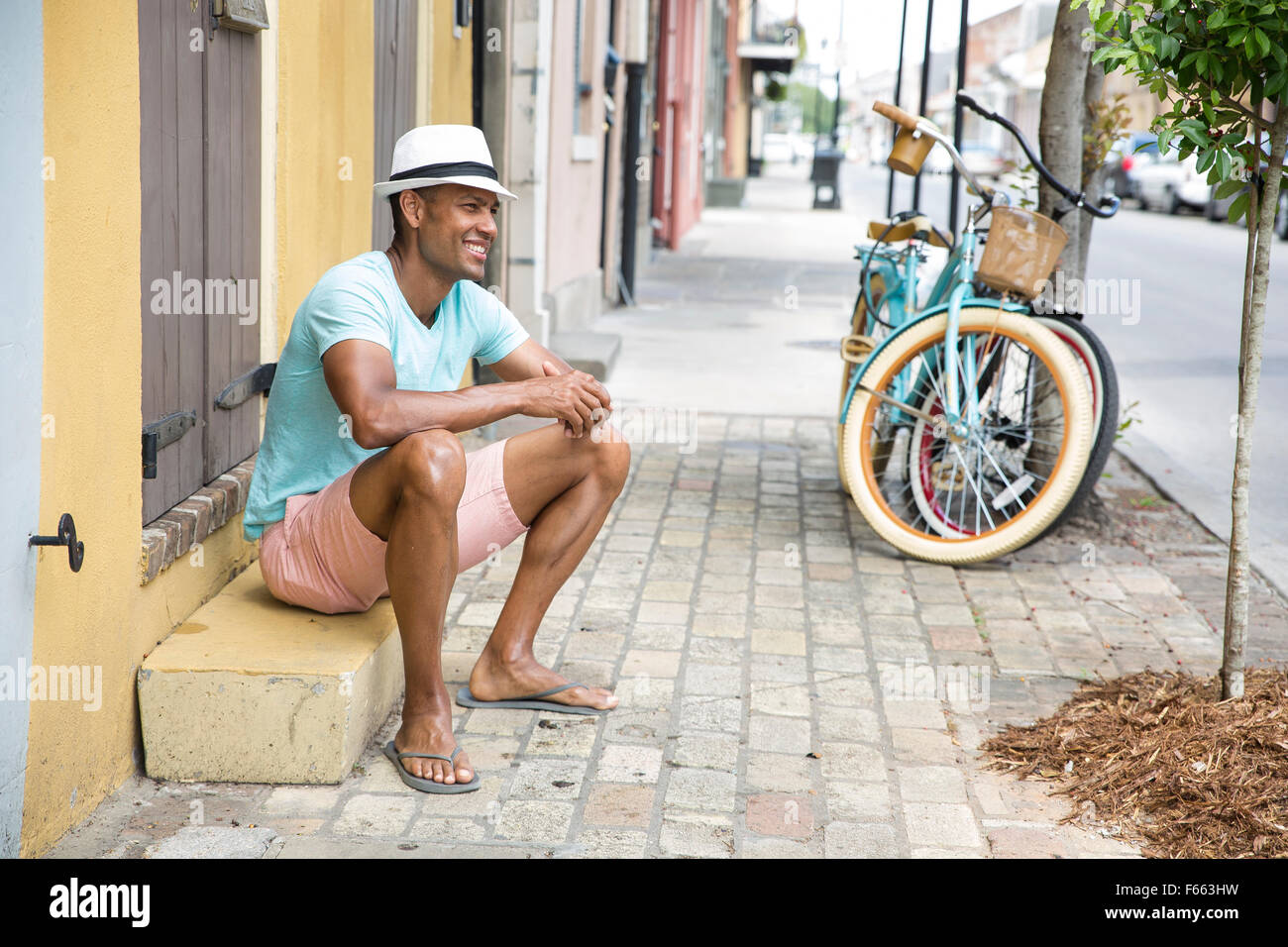 Portrait of an ethnic man smiling wearing a light blue green t-shirt and black and white fedora with a bicycle in th background. Stock Photo