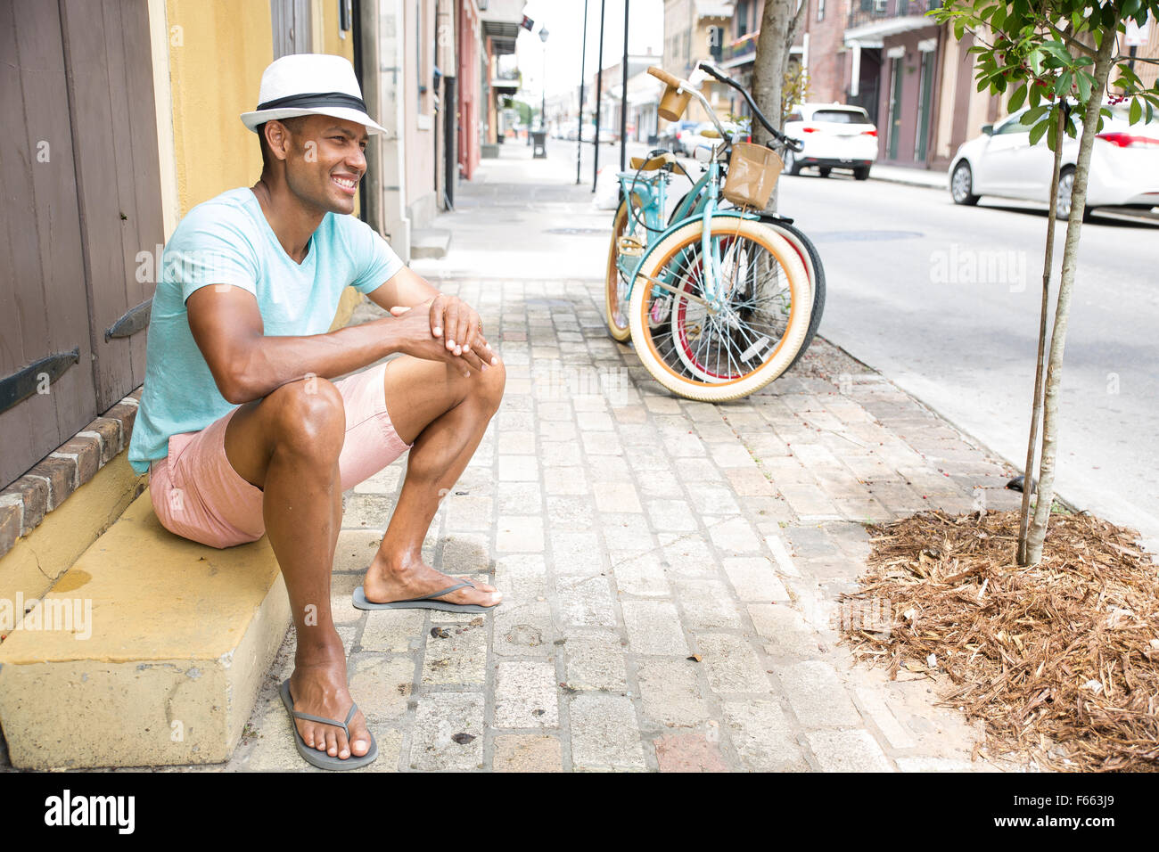 Portrait of an ethnic man smiling wearing a light blue green t-shirt and black and white fedora with a bicycle in th background. Stock Photo