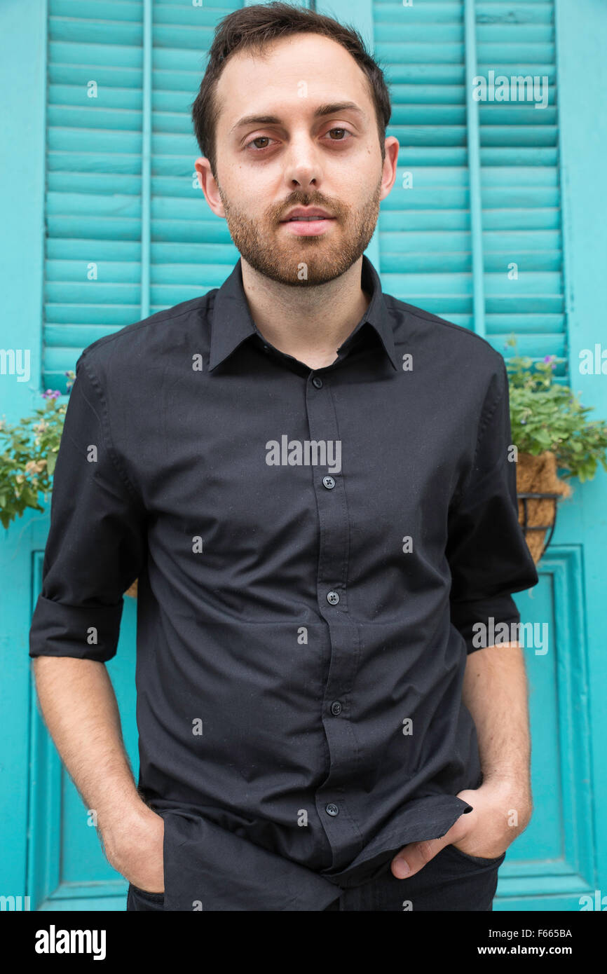 Portrait of a young man with his hands in his pockets wearing a black button up dress shirt with a greenish blue background. Stock Photo