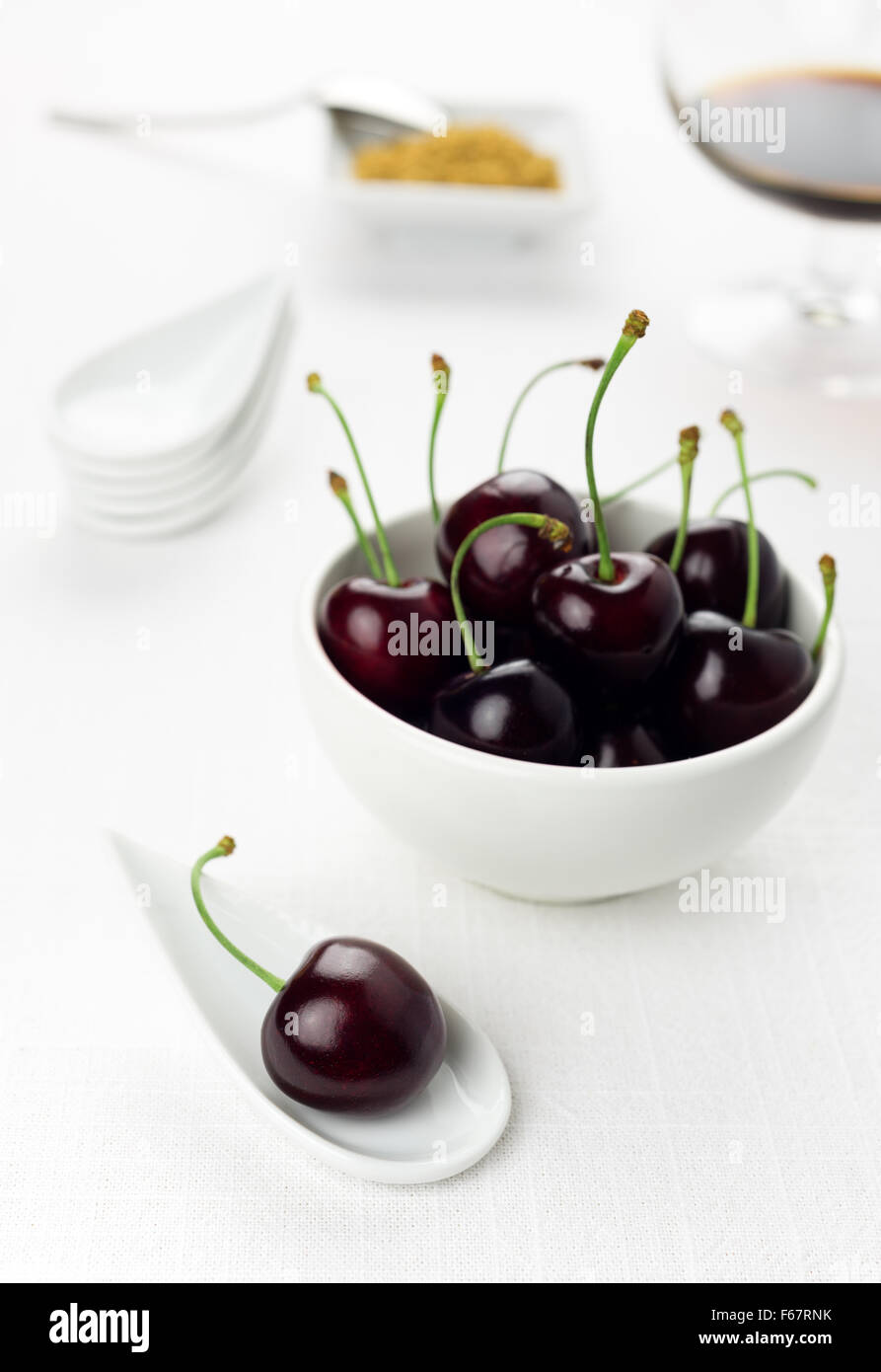 Single cherry on a white china spoon, a cherries group in a small round white bowl and a sherry glass with sugar cane on backgro Stock Photo