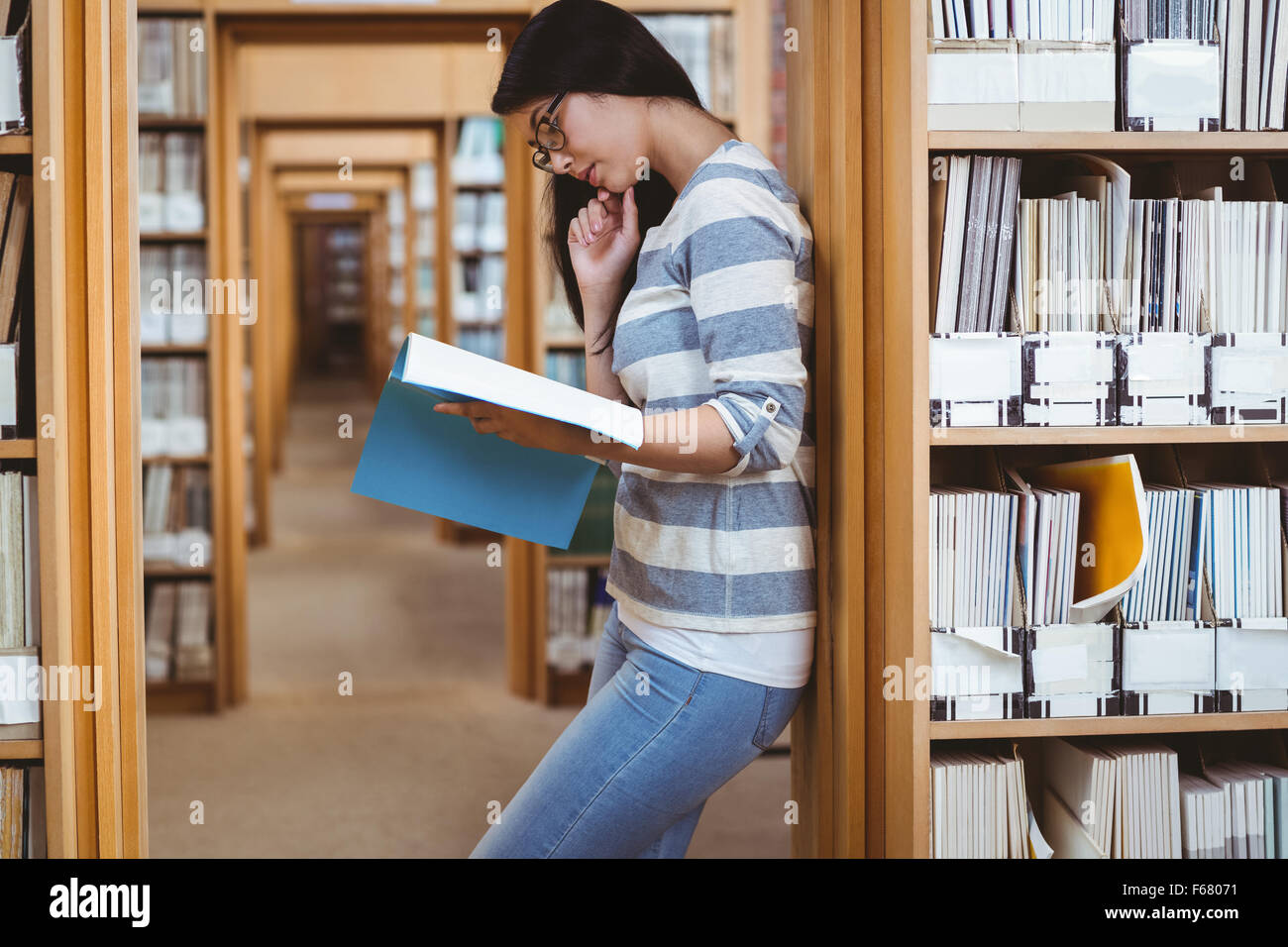 Focused student leaning against bookshelves and reading a book in library Stock Photo