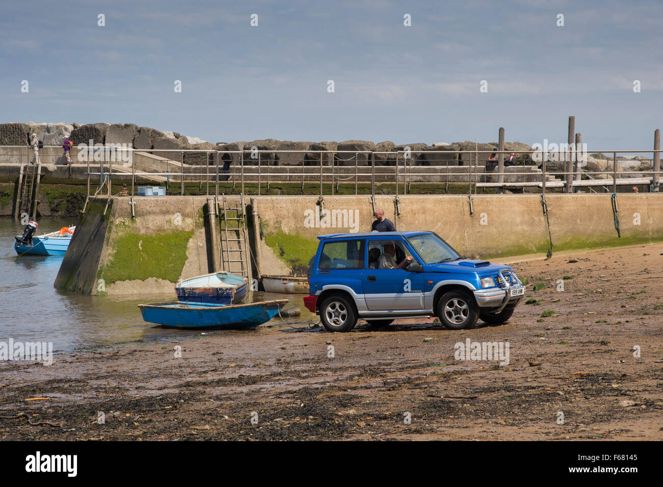 Sunny summer day with blue sky, Staithes harbour, North Yorkshire, UK - man in 4x4 Suzuki is pulling a small rowing boat out of the sea at low tide. Stock Photo
