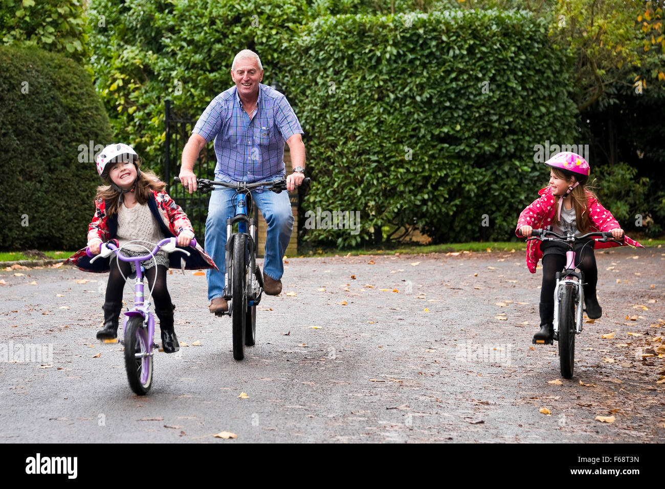 Horizontal portrait of young girls riding their bikes along the road with their granddad. Stock Photo