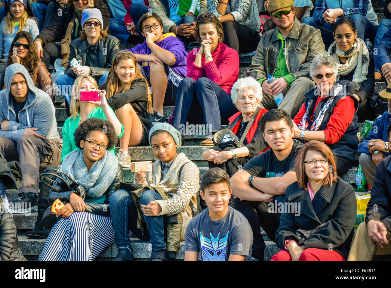 A large, diverse group of people of all ages & ethnicities sit outside in rows as an audience watching a live performance in USA melting pot city Stock Photo