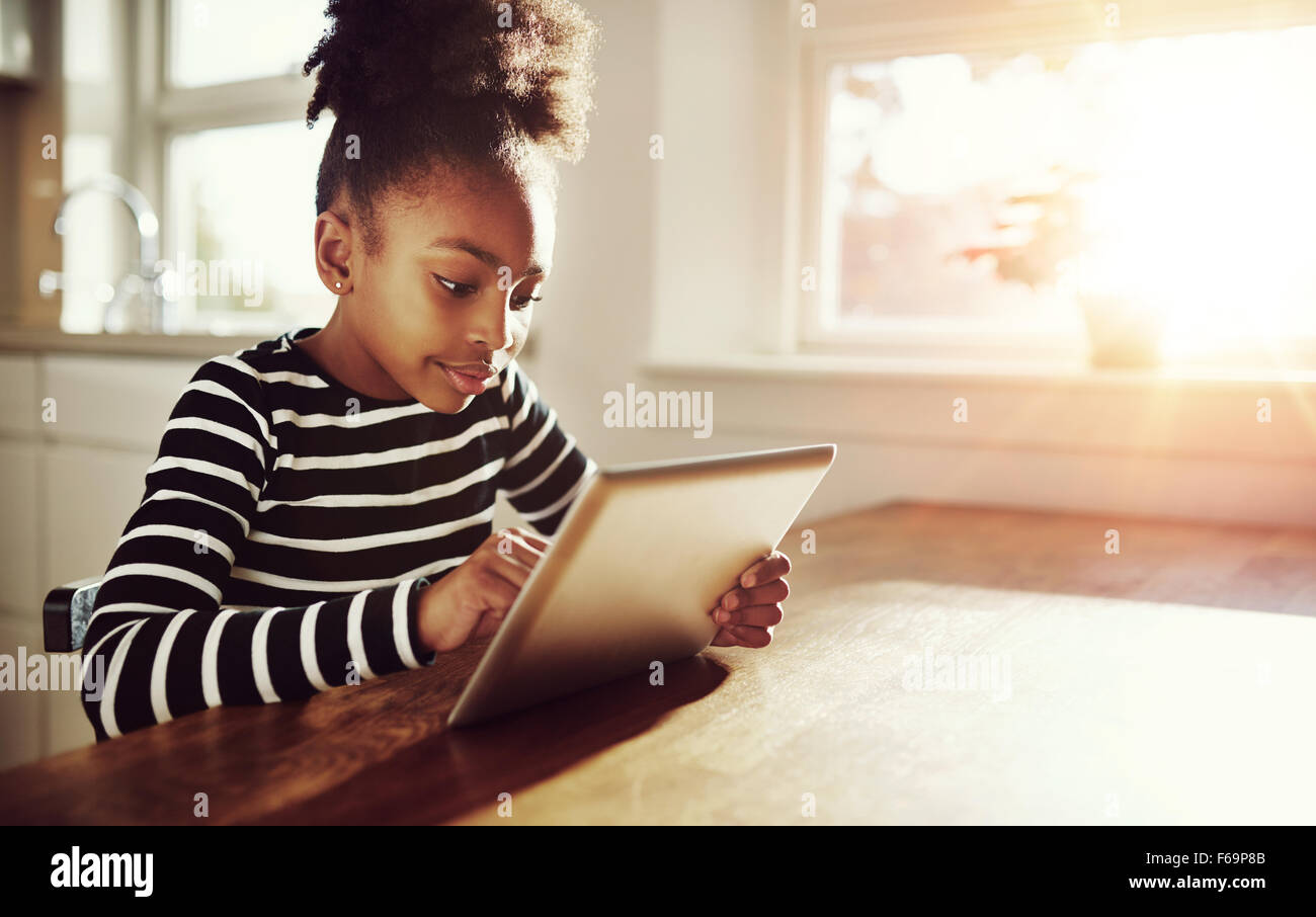 Young black girl with a fun afro hairstyle sitting at a table at home browsing the internet on a tablet computer with bright sun Stock Photo