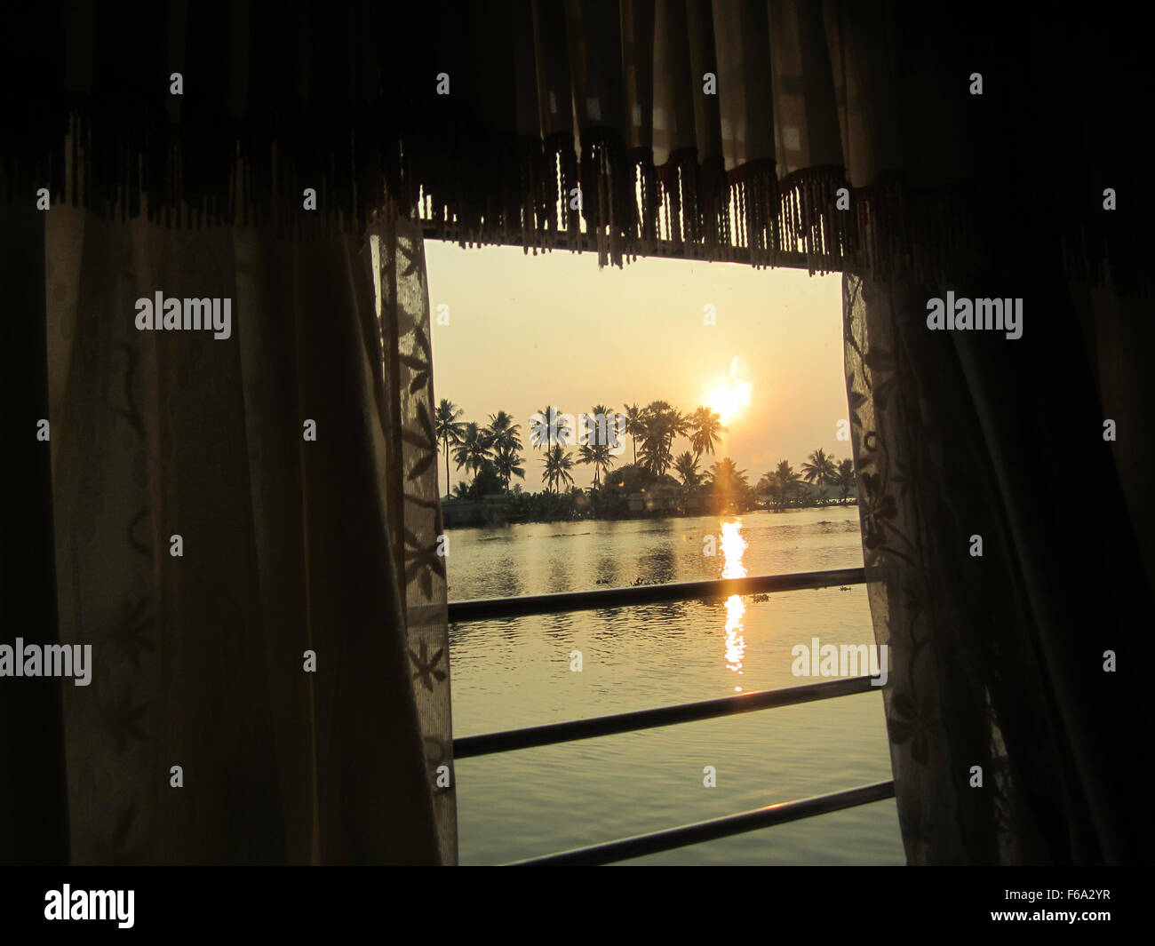 Houseboat window - sunset view showing the  calm waterway  and small cluster of palm trees framed by curtain detail Stock Photo