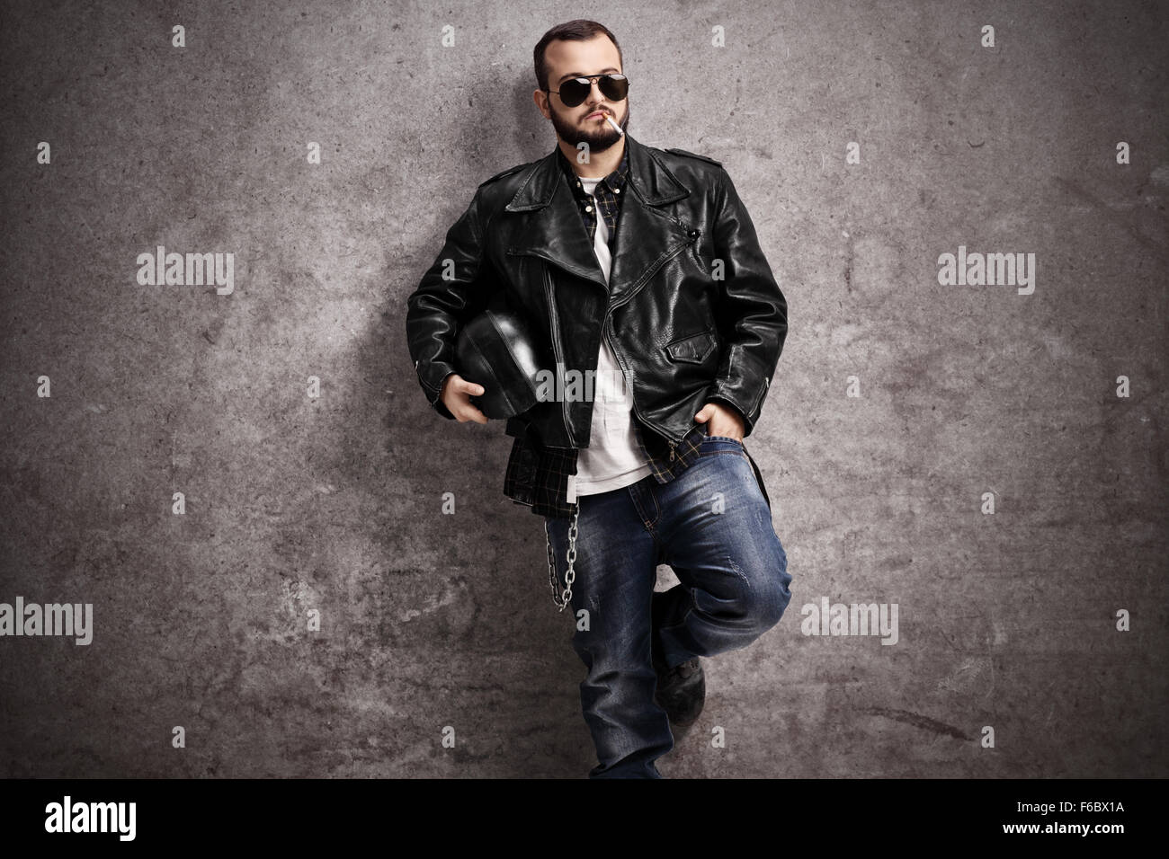 Young male biker in a leather jacket smoking a cigarette and leaning against a rusty gray wall Stock Photo