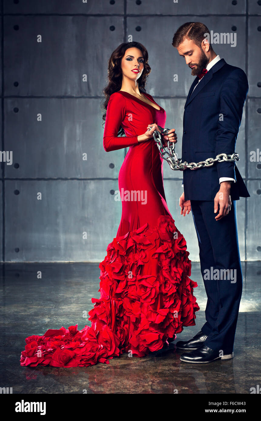 Young happy woman in red dress holding man on heavy chain. Elegant evening clothing. Stock Photo