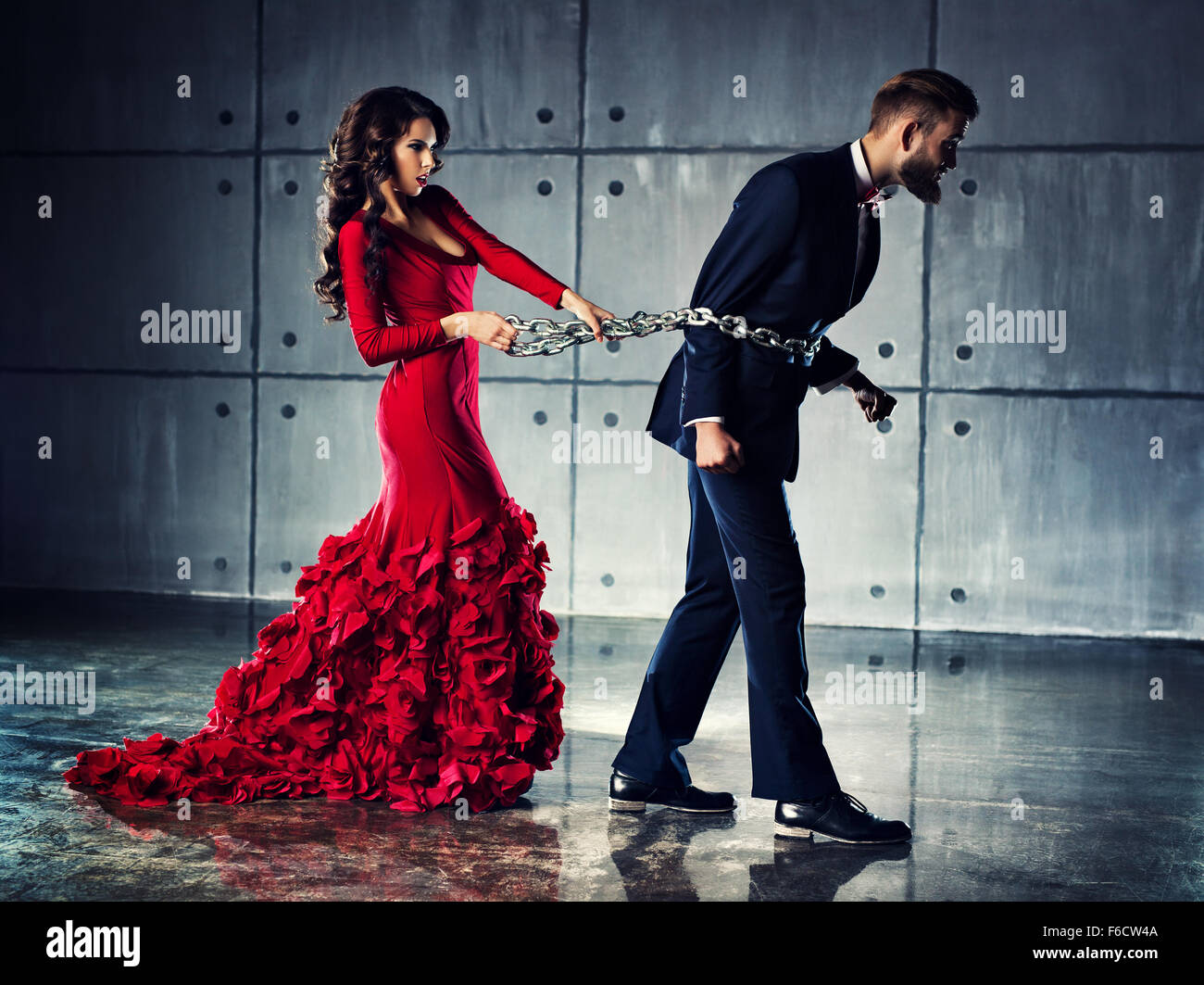 Young woman in red dress holding man on heavy chain. He tries to escape. Elegant evening clothing. Stock Photo
