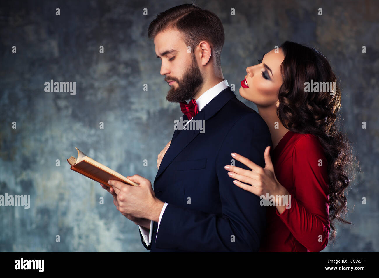 Young elegant couple in evening dress portrait. Man reading book and woman trying to attract and embrace him. Stock Photo