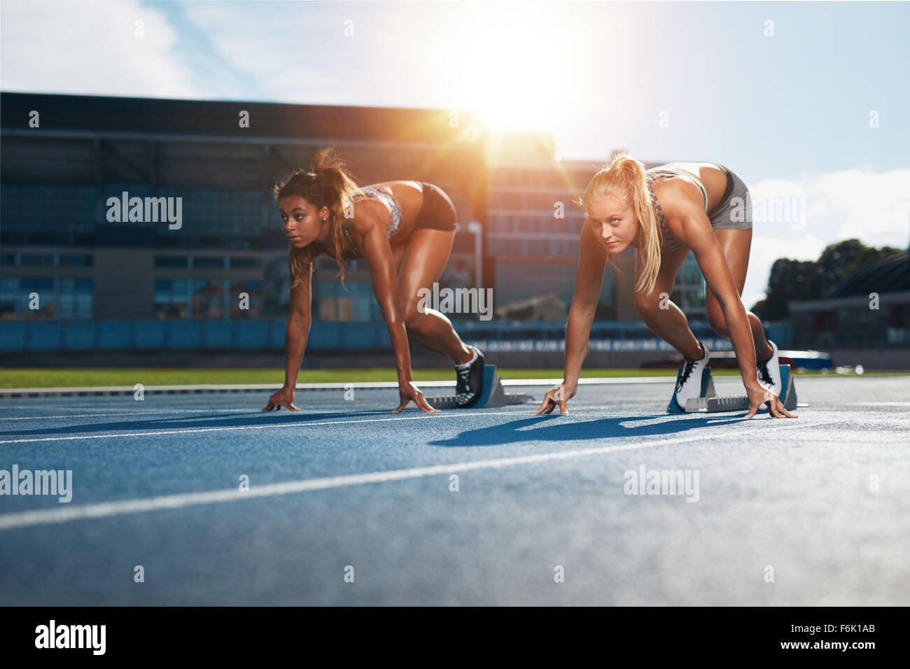 Two female athletes at starting position ready to start a race. Sprinters ready for race on racetrack with sun flare. Stock Photo