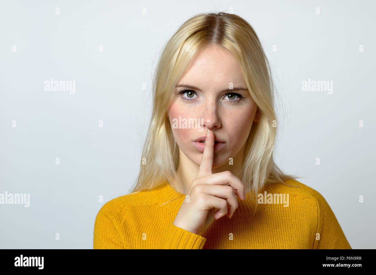 Close up Pretty Blond Woman Showing Shushing Gesture While Staring at the Camera Against Gray Background. Stock Photo