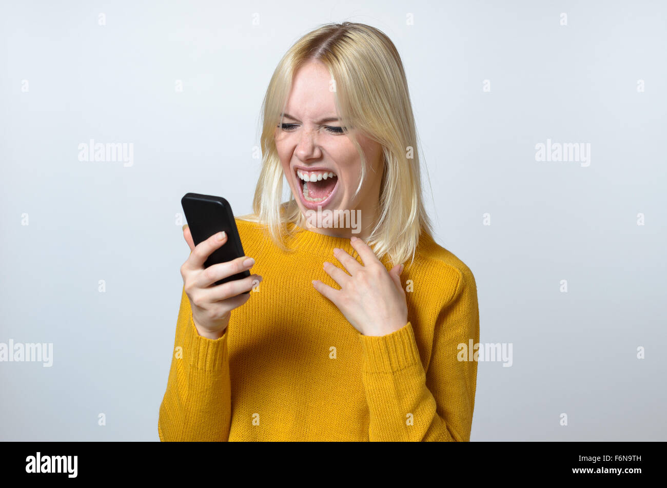 Blond Young Woman Showing Shocked Expression While Looking at her Mobile Phone Against White Background. Stock Photo