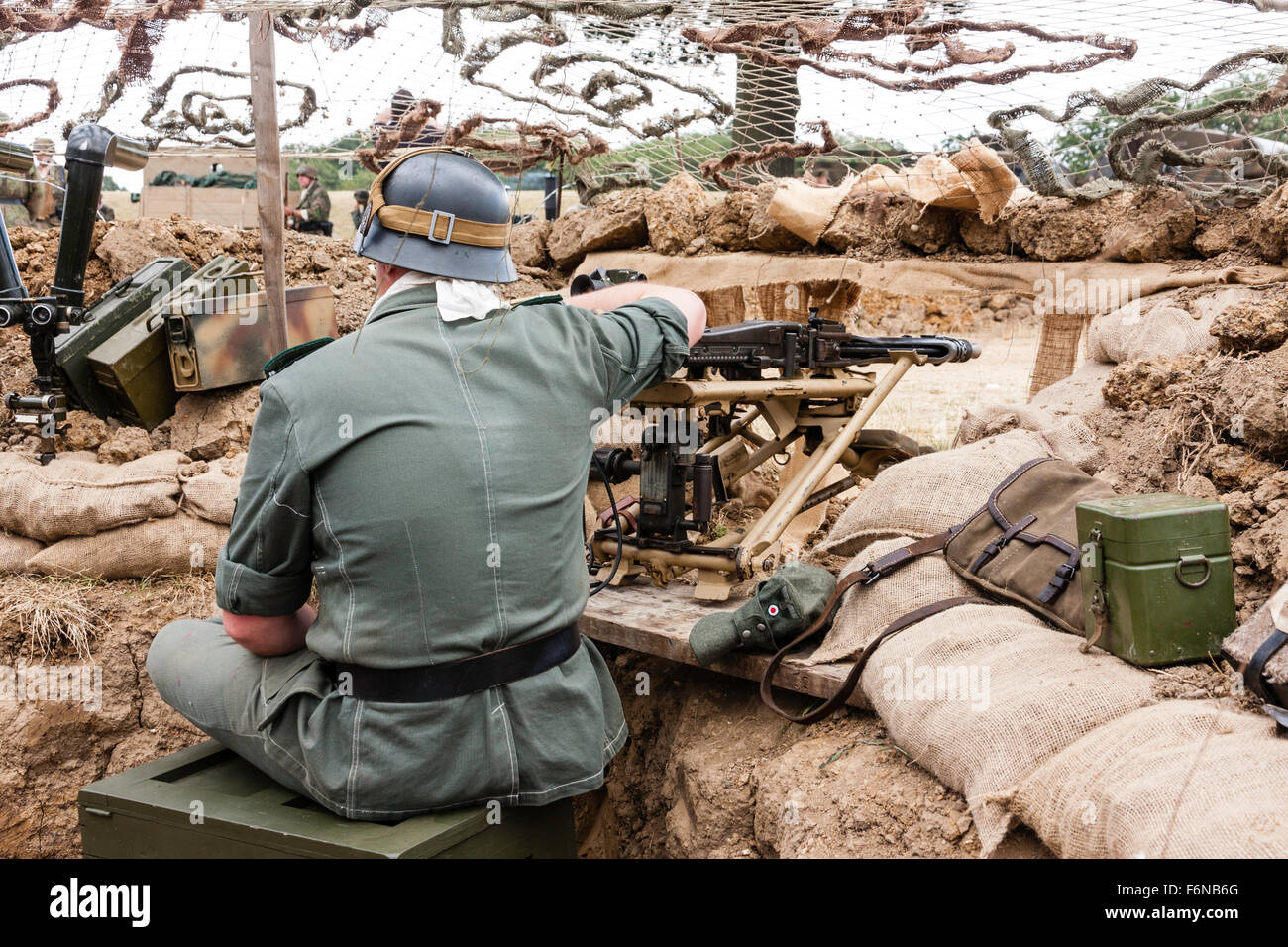 World war two re-enactment. German solider sitting, with back to viewer, in sandbagged trench manning MG42 machine gun under camouflage netting. Stock Photo