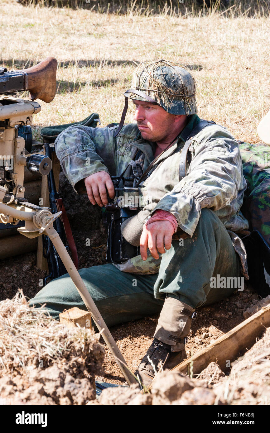 WW2 Re-enactment. German soldier in camouflage smock, sitting in trench, resting, waiting, looking weary and exhausted. Machine gun next to him. Stock Photo