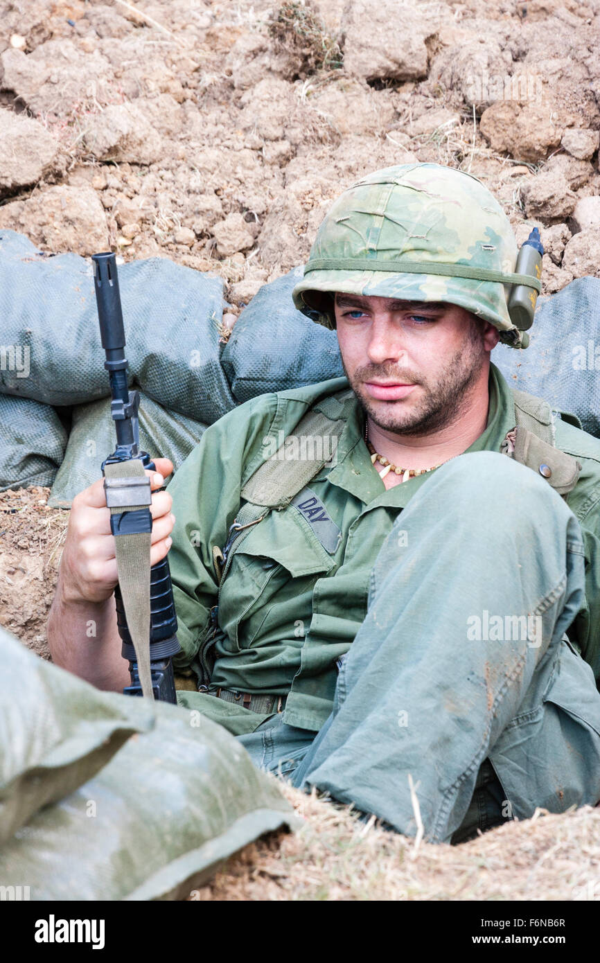 Vietnam war re-enactment. American soldier sitting in sandbag dugout, holding M16, no eye-contact but looking weary and exhausted. Facing Stock Photo