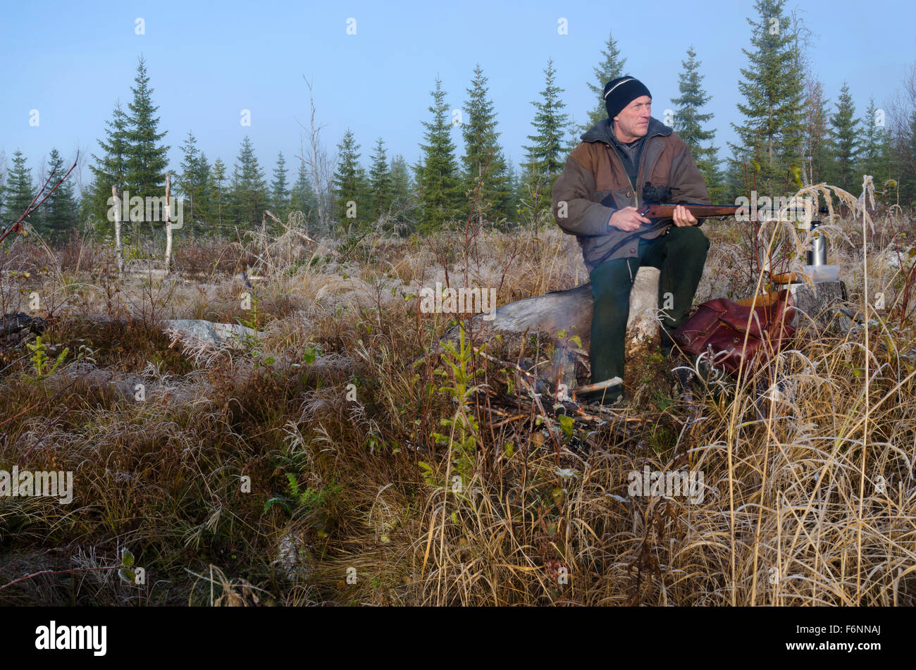 Moose hunter sitting on a stump with a litle fire in front holding his rifle pointing to the left. Stock Photo