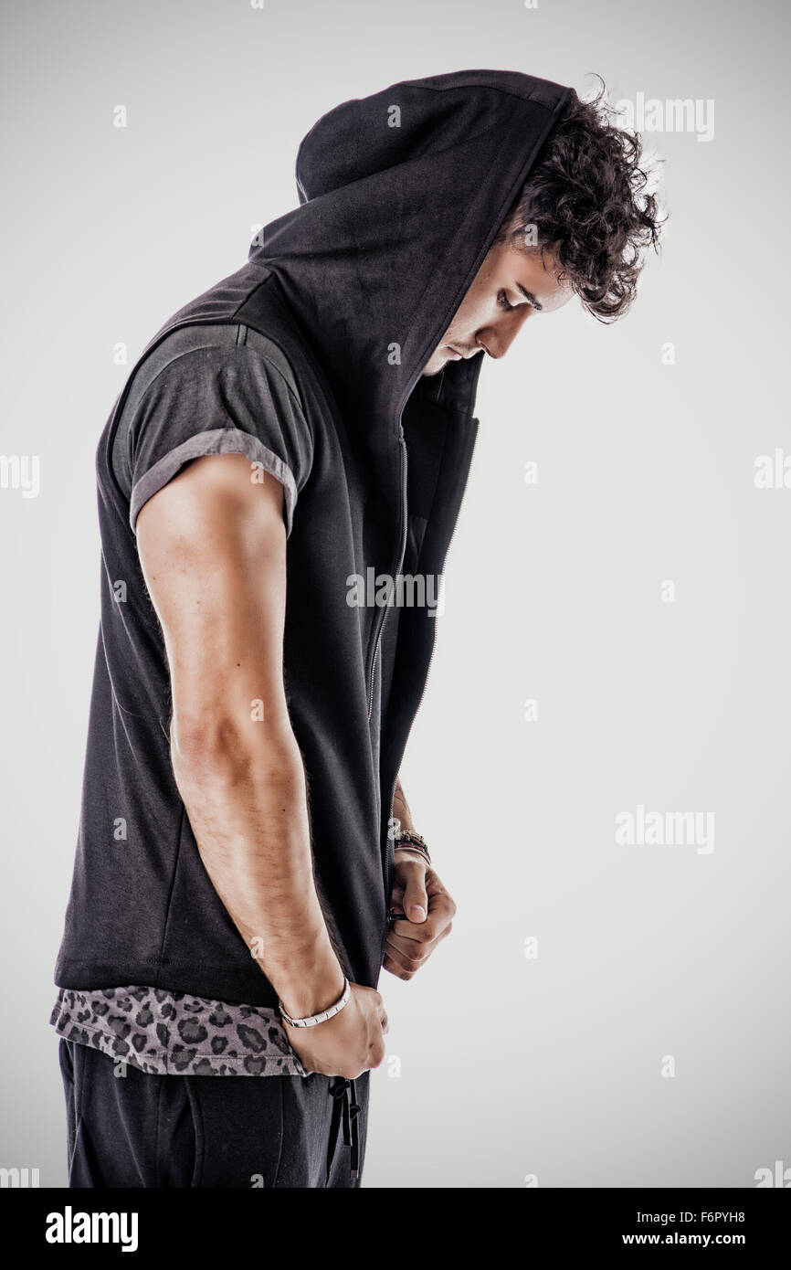 Side view of handsome tough young man in dark hooded t-shirt on light background, zipping up the shirt, looking down Stock Photo