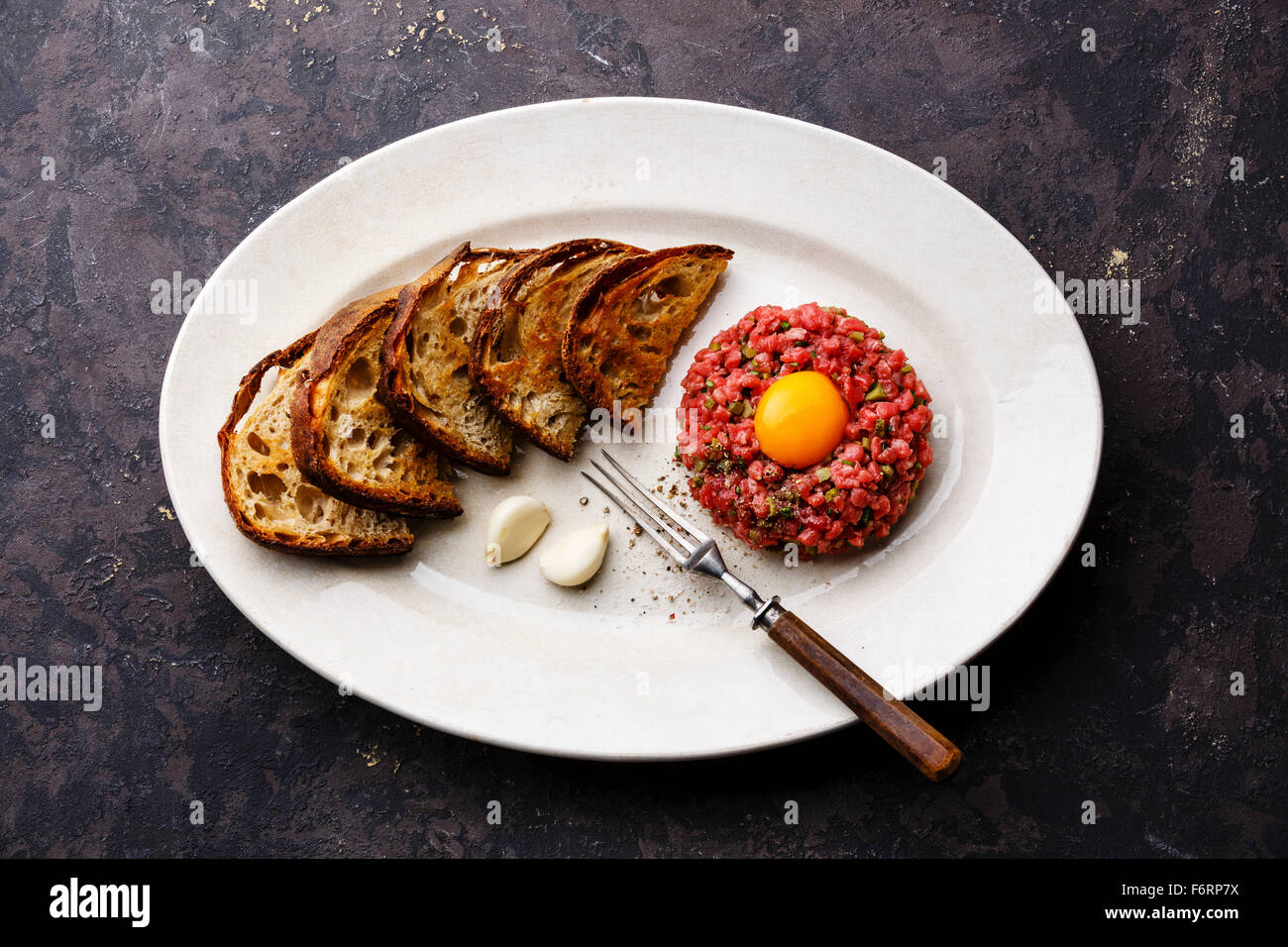 Beef tartare with pickled cucumber and toasts on white plate on dark background Stock Photo