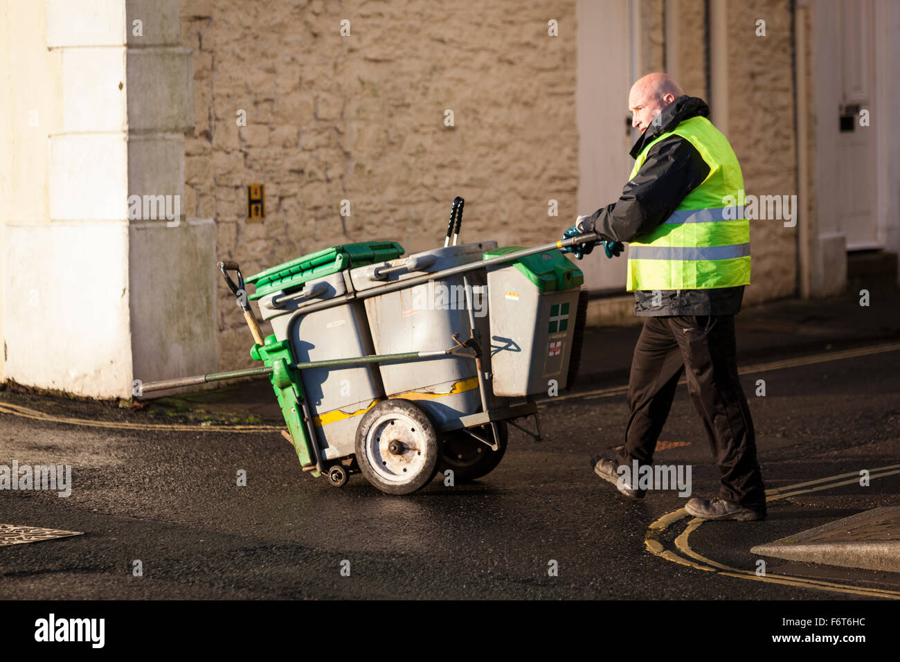 A street sweeper wearing a yellow hi-viz jacket, pushes his cleaning equipment trolley across a damp road in Dartmouth, Devon. Stock Photo