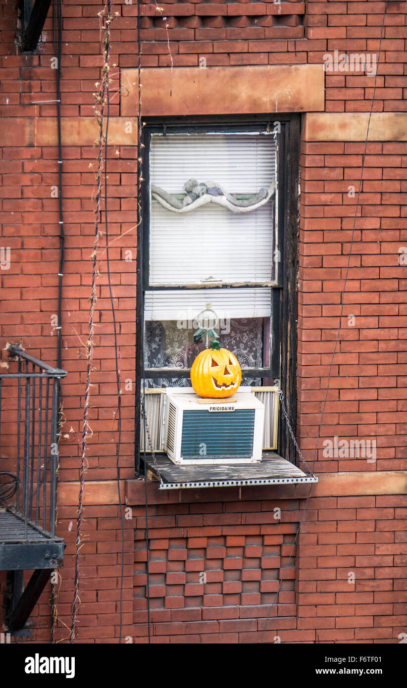 bright orange ceramic version hand carved Halloween pumpkin sitting on air conditioner outside window brick tenement building Stock Photo
