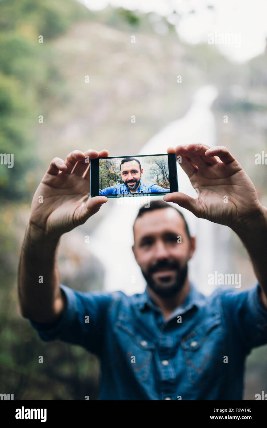 Bearded man taking a selfie with smartphone in front of a waterfall ...
