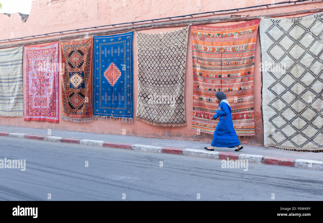 Moroccan woman walking past carpets hanging on wall, Marrakech, Morocco Stock Photo