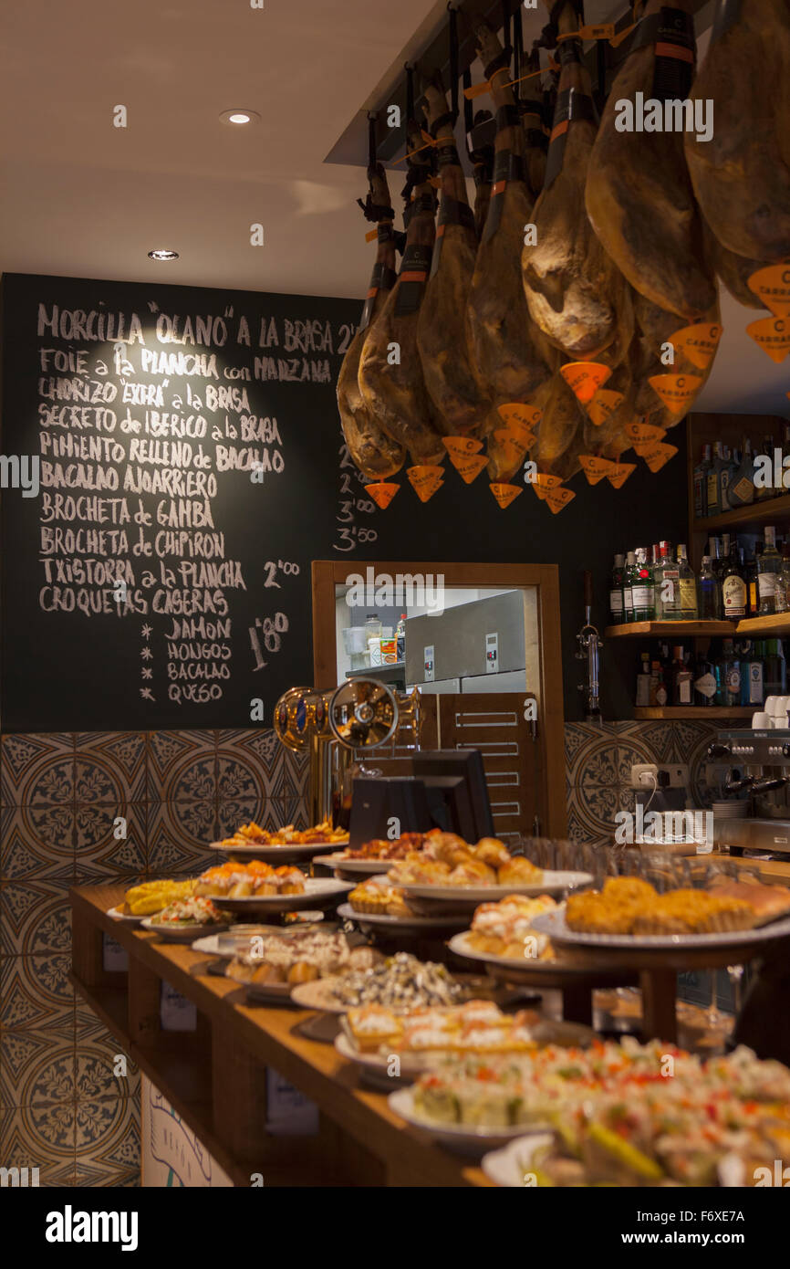 Pinxos on a bar counter, hams hanging from the ceiling and a menu written on a blackboard in a bar called Portaletas in the old town Stock Photo