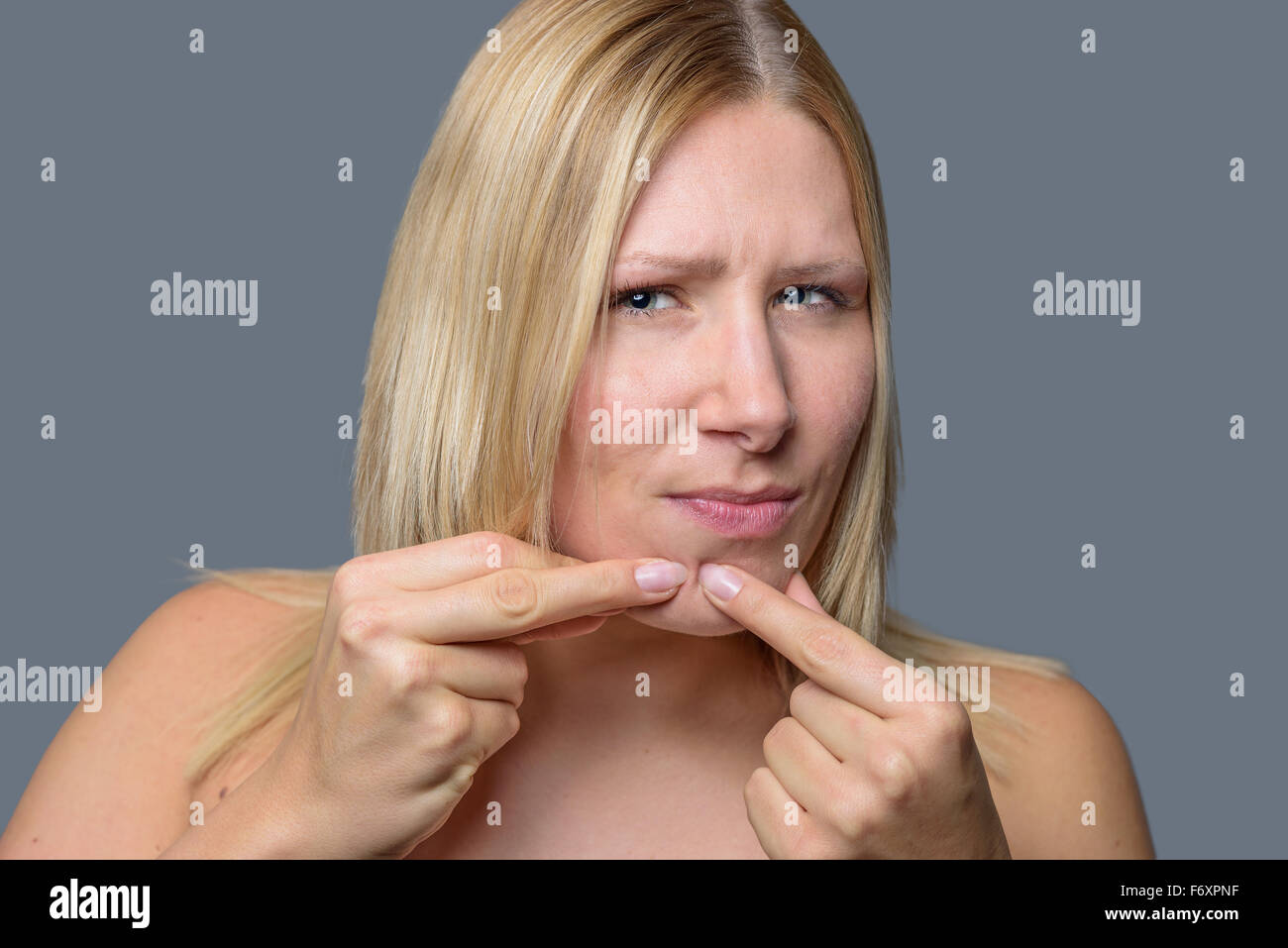 Woman squeezing a pimple, spot zit or blackhead on her chin with her fingers, close up of her face and hands over grey Stock Photo