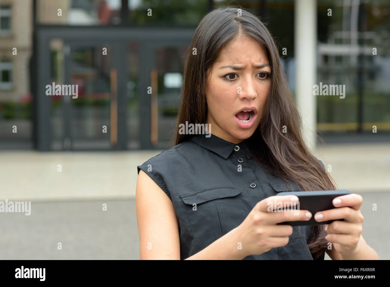 Young woman reacting in horror to an sms, text message or email on her mobile phone as she stands outdoors in a quiet urban stre Stock Photo