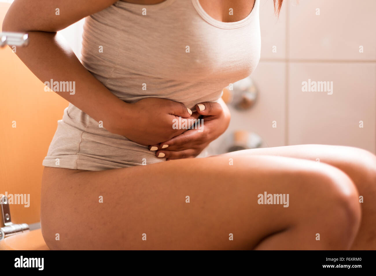 Close up Sick Young Woman Sitting on the Toilet in the Bathroom While Holding her Painful Stomach Stock Photo
