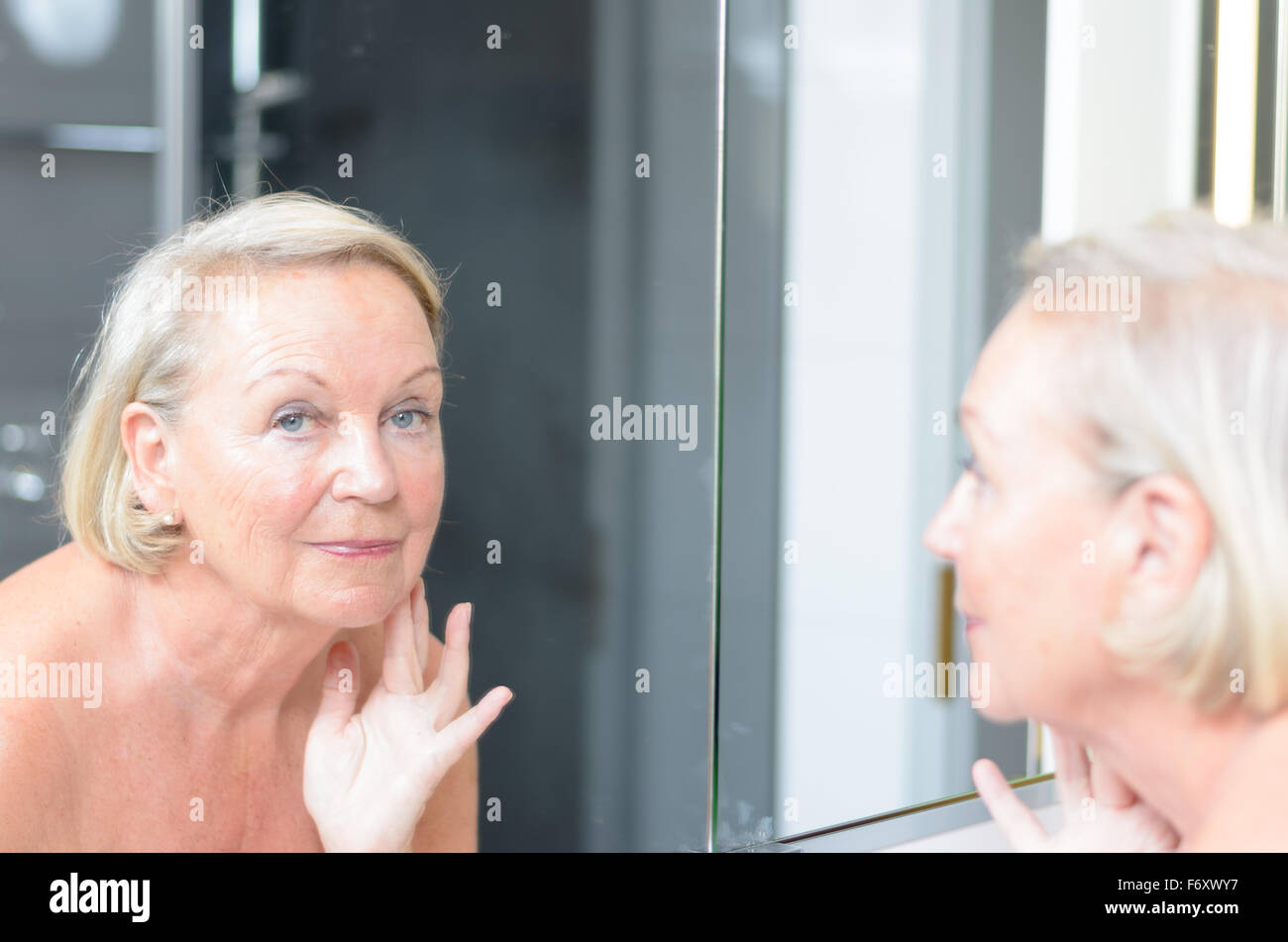 Senior lady checking her skin in the mirror leaning forwards for a closer look as she hold a finger to her forehead Stock Photo