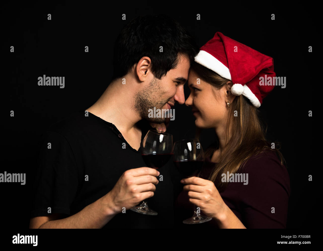Young Christmas couple, wearing red hut, holding wine glasses, on black background, touching noses Stock Photo
