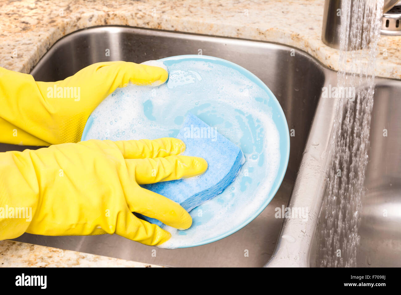 A person washes dinner plates using soapy detergent, a sponge and protective gloves then rinses them before drying Stock Photo