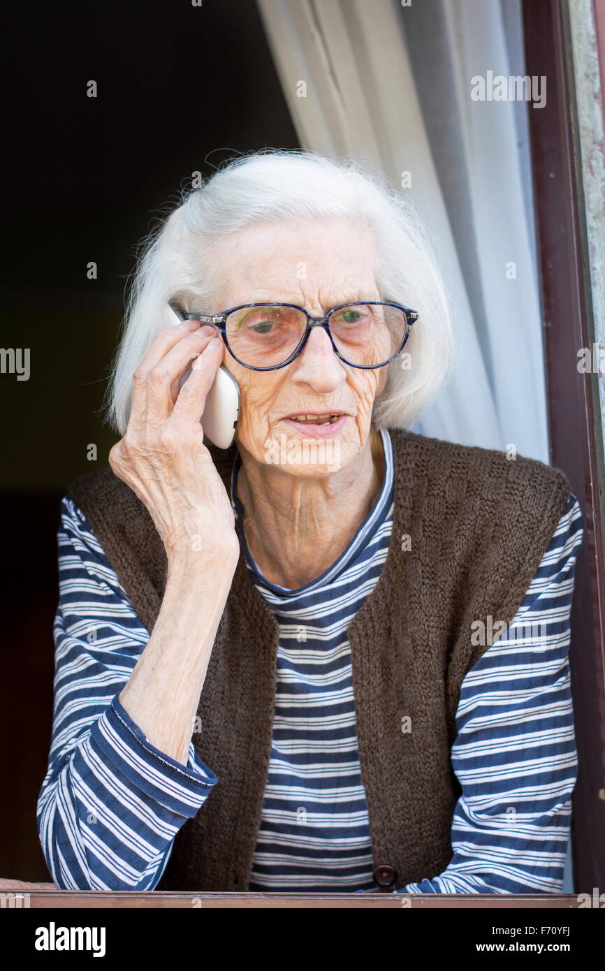 Old grandma talking on the phone while standing on her home window Stock Photo