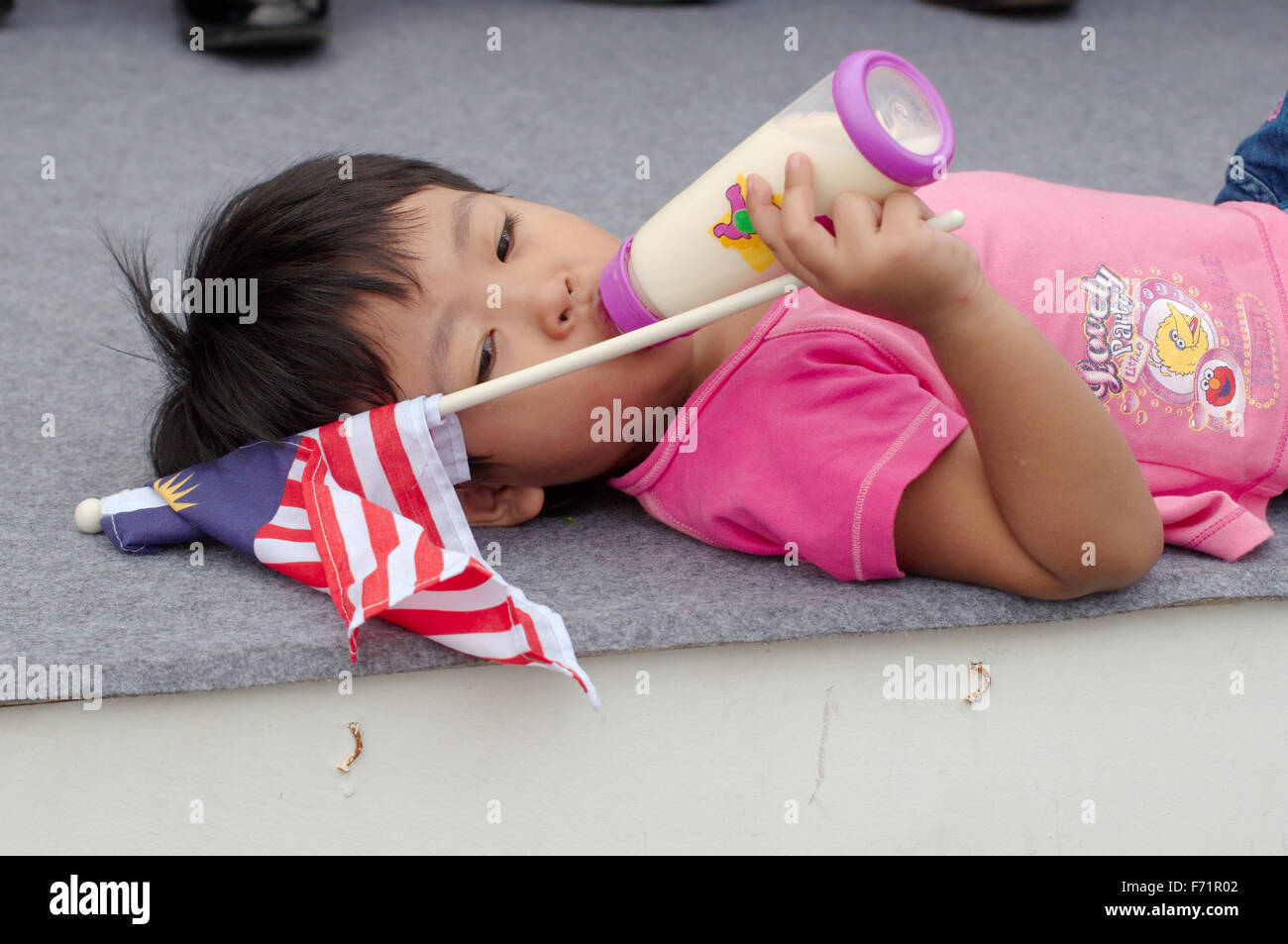 The little Malaysian girl lies on the scene and drinking milk from a nipple, holding a Malaysian flag, Kuala Lumpur, Malaysia Stock Photo