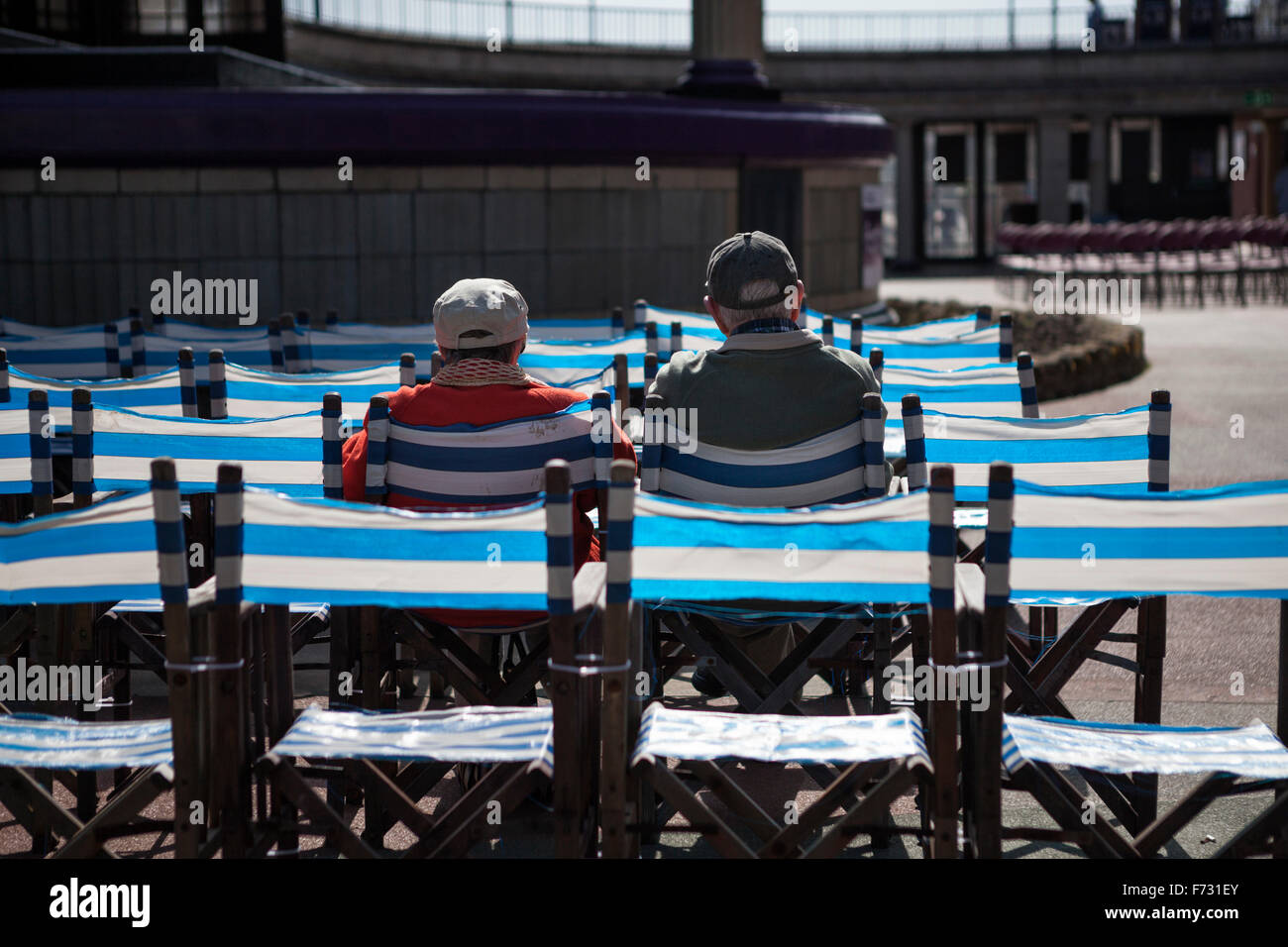 Couple sitting in blue and white striped chairs outdoors in the sunshine at Eastbourne bandstand, Eastbourne, East Sussex, England, UK Stock Photo