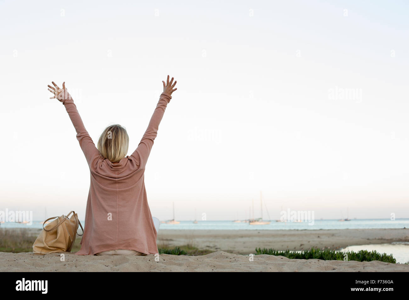 Blond woman sitting on a sandy beach, arms raised. Stock Photo