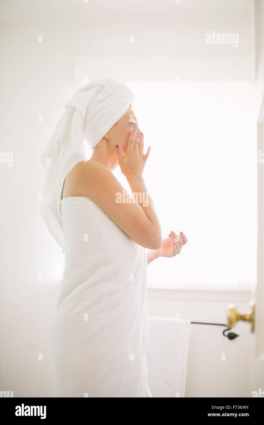 Woman wrapped in a white towel standing in a bathroom, applying cream to her face. Stock Photo