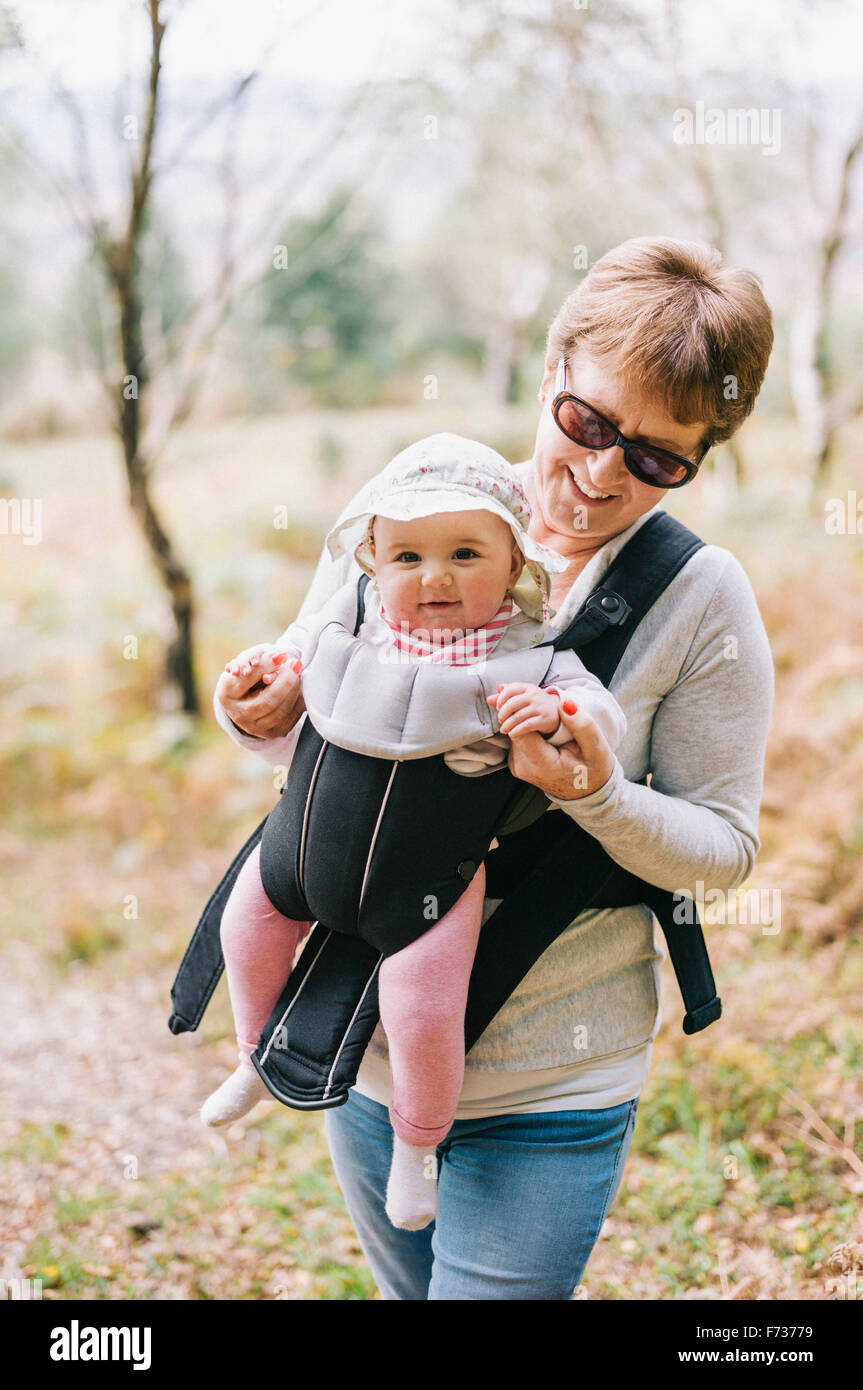 A woman wearing sunglasses carrying a baby wearing a white sunhat in a front baby sling. Stock Photo