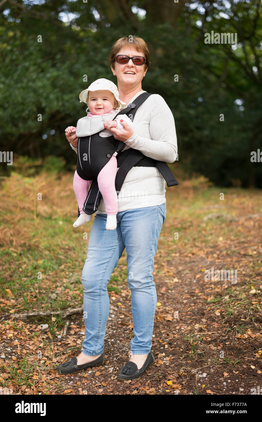 A woman wearing sunglasses carrying a baby wearing a white sunhat in a front baby sling. Stock Photo