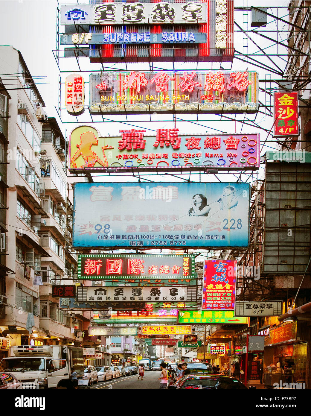 This is a magnificent view of a street in Kowloon, Hong Kong with high-rise buildings and nicely park vehicles on its both side, there are numerous glowing signboards above the street spotted throughout the length of the street. Stock Photo