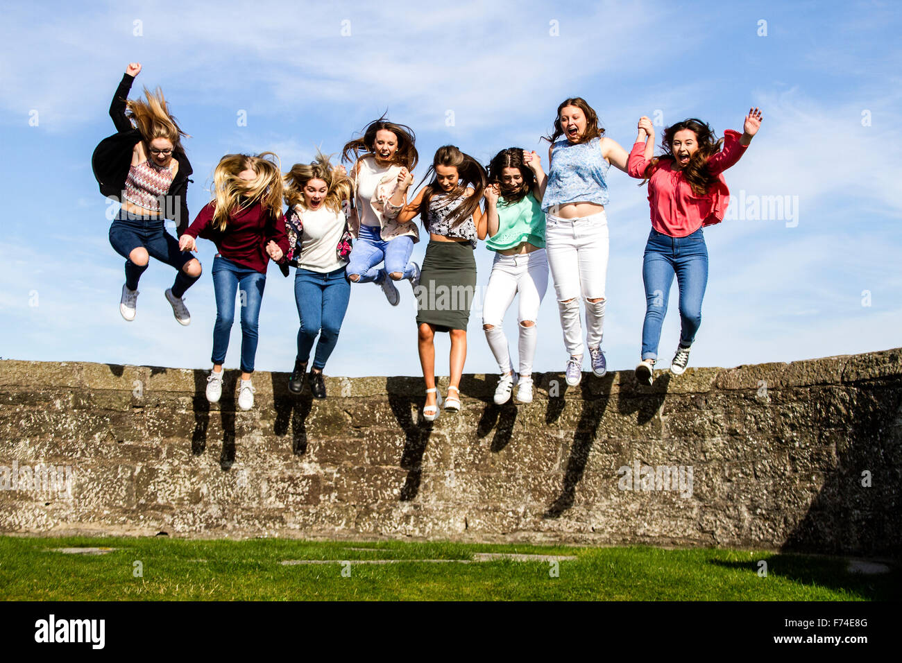 Eight teenage girls having fun holding hands and jumping off the Broughty Ferry Castle wall on a sunny day in Dundee, UK Stock Photo