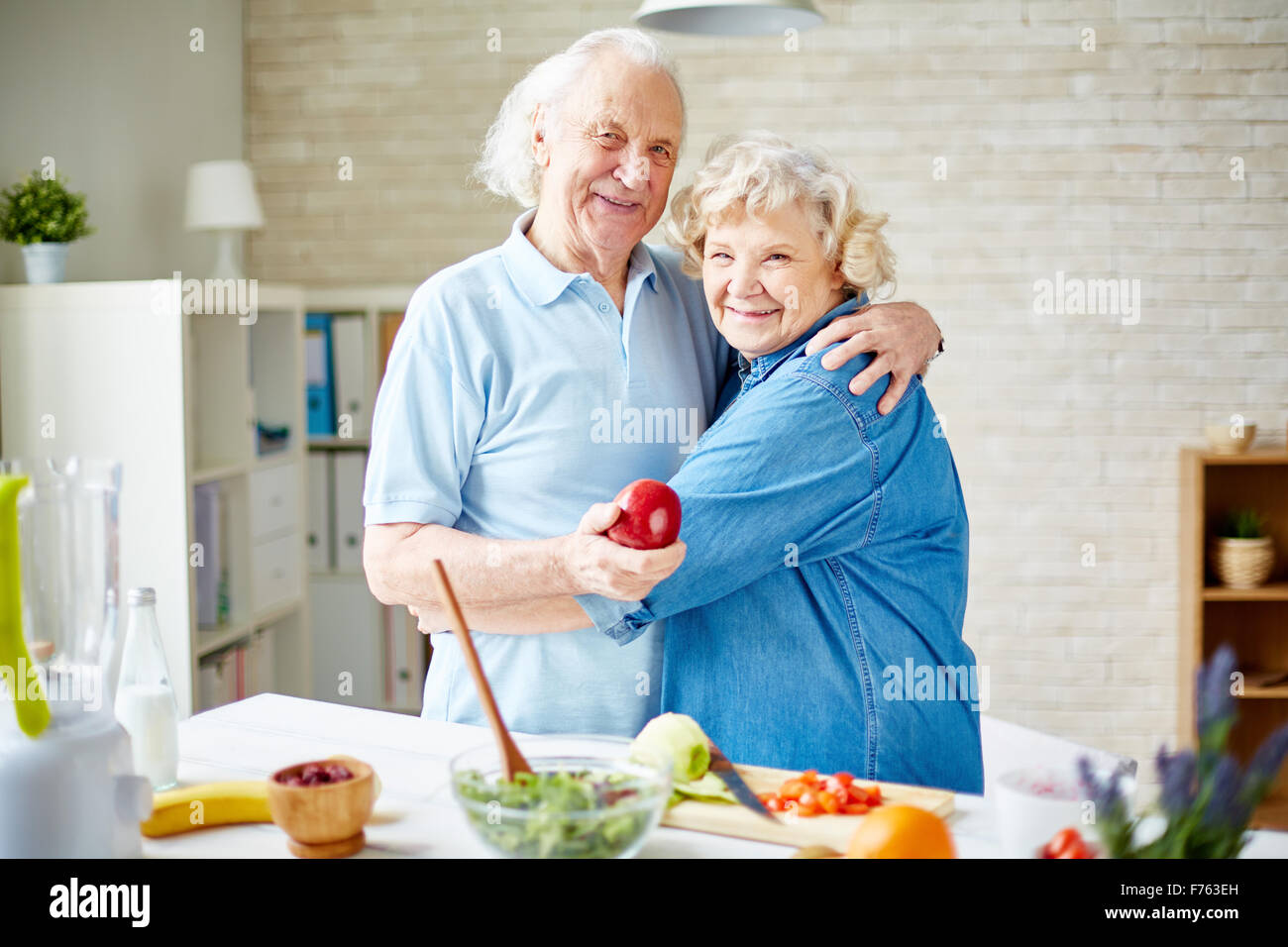 Happy senior husband and wife in embrace looking at camera in the kitchen Stock Photo