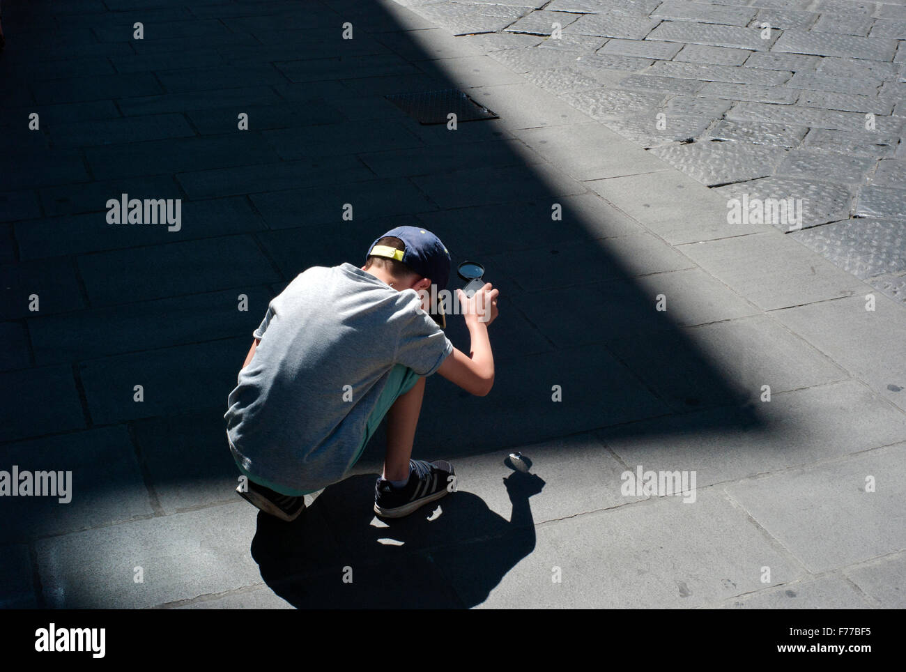 boy crouches on street Burning Paper Using Magnifying Glass Stock Photo