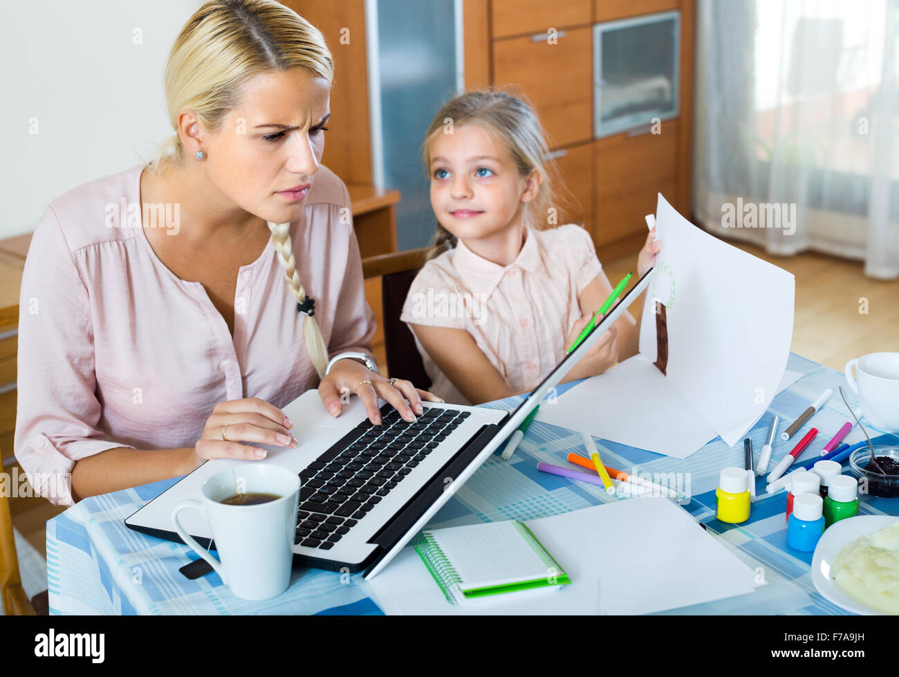 Tired young woman irritated as daughter diverts her from work at home Stock Photo