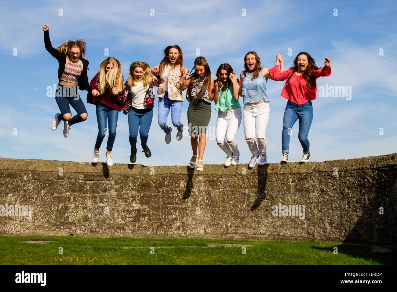 Eight teenage girls having fun holding hands and jumping off the Broughty Ferry Castle wall on a warm sunny day in Dundee, UK, Stock Photo